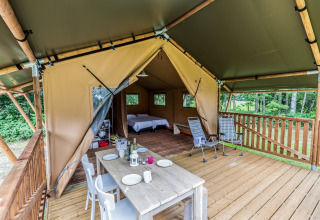 Tente safari avec terrasse en bois, table à manger extérieure, fauteuils et vue sur la forêt.