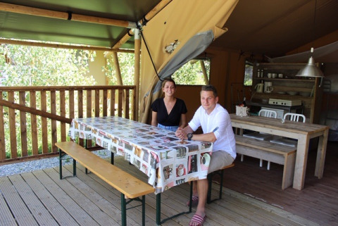 Twee mensen zitten aan een tafel in een safaritent met houten meubels en een keuken op de achtergrond.
