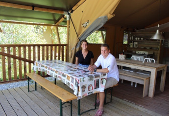 Two people sit at a table inside a safari tent, with wooden furniture and a kitchen area in view.