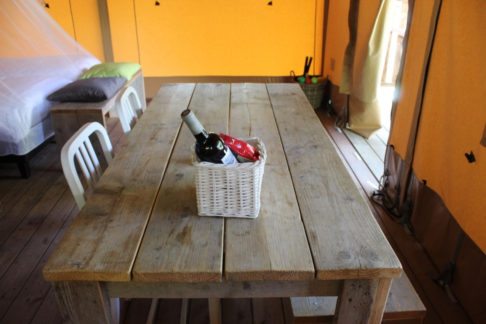 Rustieke houten tafel in een safaritent met mand met wijn en snacks, bed en stoelen op de achtergrond.