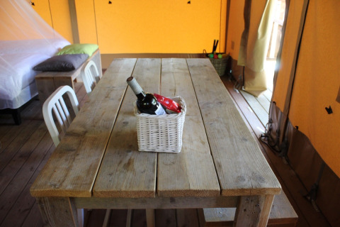 Rustikaler Holztisch in einem Safarizelt mit einem Korb mit Wein und Snacks, Bett und Stühle sichtbar.