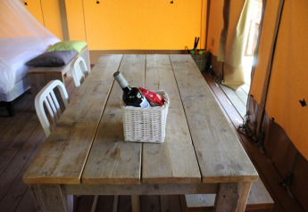 Table en bois rustique dans une tente safari, panier avec vin et snacks, lit et chaises en arrière-plan.