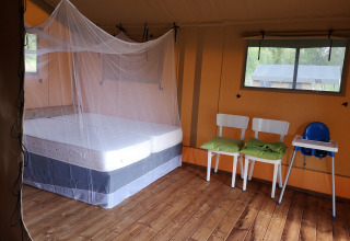 Interno di tenda safari con letto matrimoniale sotto zanzariera, due sedie e seggiolone su legno.