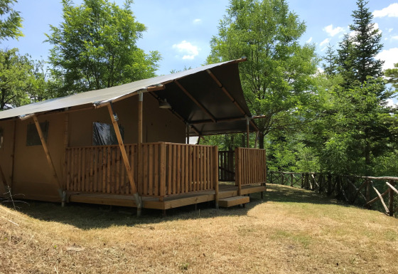 Tenda safari con servizi igienici su prato erboso, circondata da alberi verdi e natura rigogliosa.