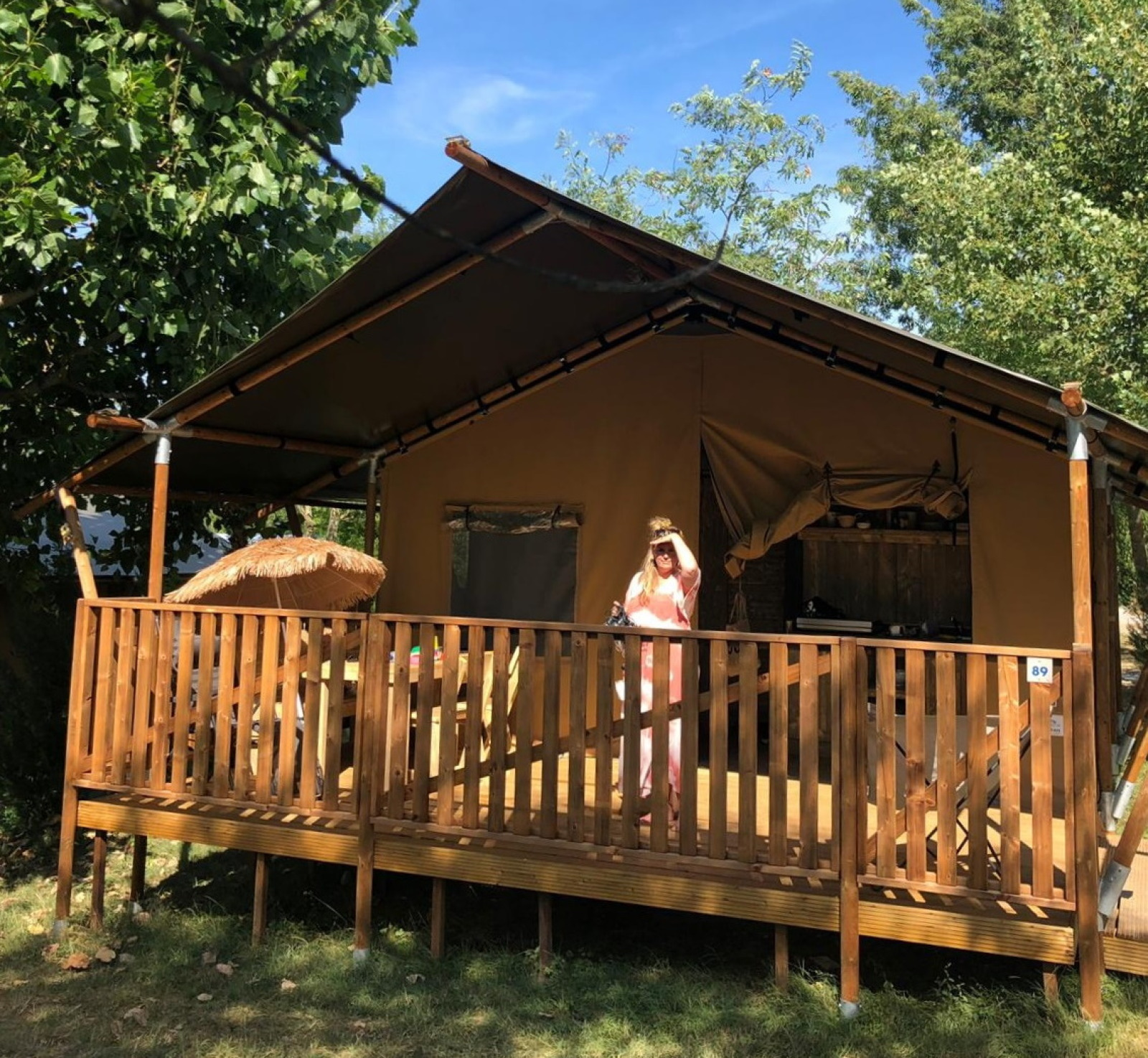 Een persoon staat op de veranda van een safaritent met sanitair, omgeven door bomen en zonlicht.