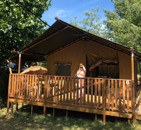 Una persona está en la terraza de una tienda safari con baño privado, rodeada de árboles y luz solar.