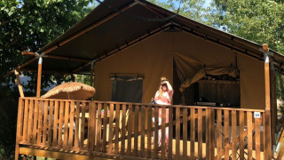 A person stands on the porch of a safari tent with private bathroom, surrounded by trees and sunlight.