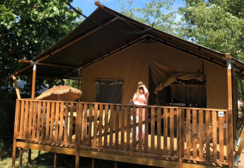 Une personne se tient sur la terrasse d'une tente safari avec sanitaires, entourée par des arbres et du soleil.