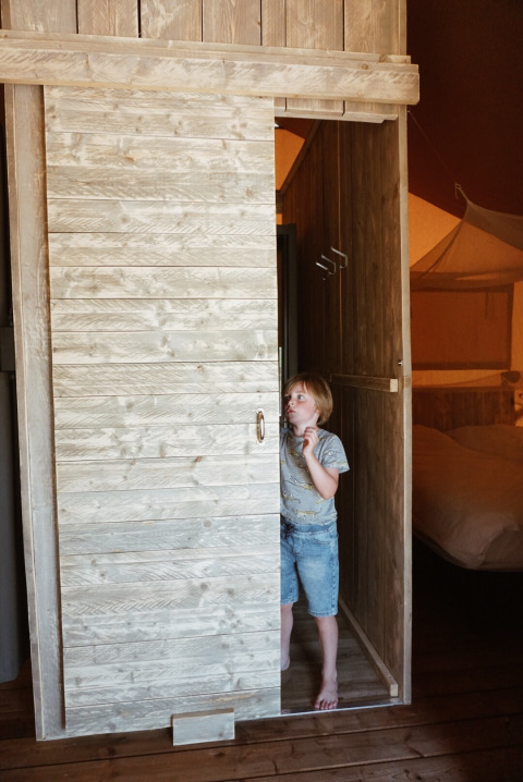 Bambino con abiti casual nella cabina di legno con porta scorrevole in una tenda safari al Camping Saint Amand, Francia.