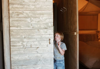 Enfant en tenue décontractée debout dans une cabane en bois à porte coulissante sous tente safari au Camping Saint Amand, France.