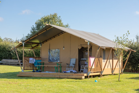 Tente safari avec terrasse en bois sur pelouse verte, chaises extérieures et table sous ciel bleu clair