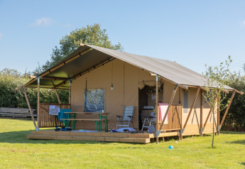 Safaritent met houten veranda op groen gras, buitenstoelen en tafel onder een heldere blauwe lucht