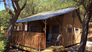 Safari-Zelt mit Veranda und Sanitäreinrichtungen im Wald, tagsüber bei blauem Himmel aufgenommen.