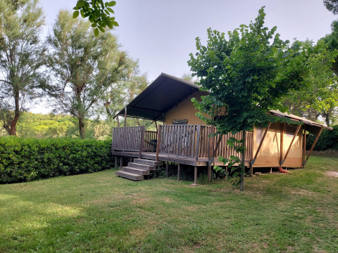 Tienda safari con instalaciones sanitarias, porche de madera y rodeada de árboles y césped verde.