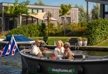 Paar genießt eine Bootsfahrt mit Hund vor modernen Glamping-Unterkünften und grünen Gärten.