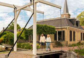 Koppel met hond wandelt over brug bij glampingverblijf, idyllisch huisje en groene omgeving erachter.