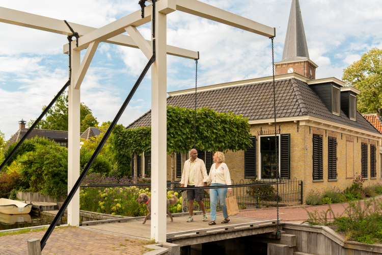 Couple with dog walking on a bridge at a glamping accommodation, with a quaint house and greenery nearby.