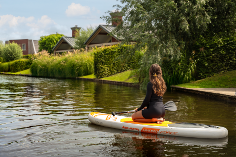 Vrouw peddelt op een opblaasbaar paddleboard langs moderne glamping huisjes aan het water en groen.