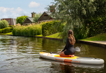 Vrouw op een opblaasbaar paddleboard bij moderne glamping huisjes tussen het groen langs het water.