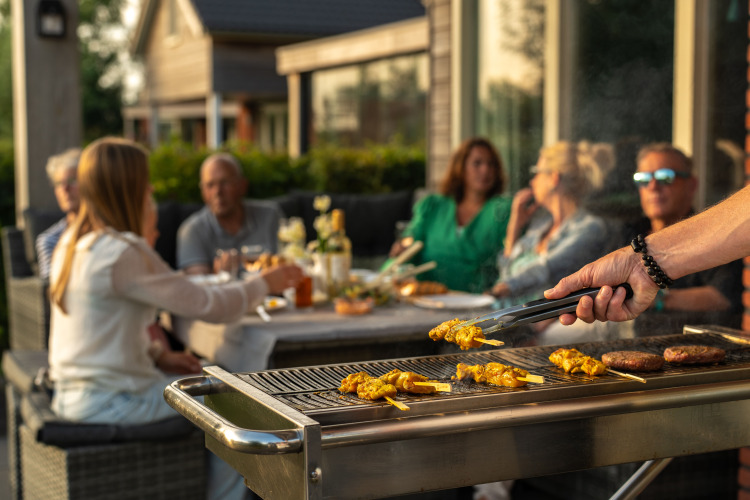 Barbacoa al aire libre en una cabaña con gente reunida en la mesa mientras alguien cocina pinchos y hamburguesas.