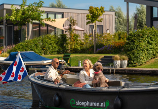 Ein Paar und ihr Hund fahren gemütlich im Boot vor einer modernen Lodge am Wasser mit viel Grün.