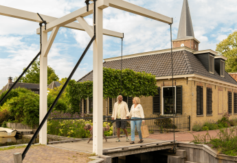 Een koppel wandelt met hun hond over een witte brug voor een sfeervolle lodge met zwart dak en groene tuin.