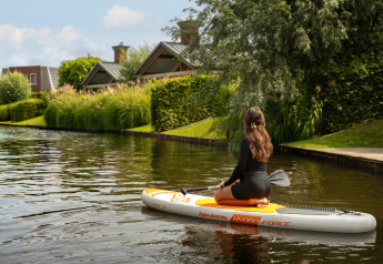 Kvinde på knæ på et paddleboard nær en lodge ved kanalen, omgivet af grønne buske og træer.
