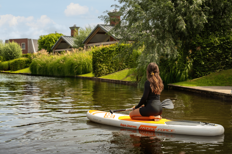 Mujer arrodillada sobre una tabla de paddle en un canal junto a una cabaña, rodeada de vegetación.