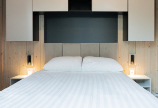 Moderne lodge-værelse med en nydelig seng, hvide puder, træpaneler og minimalistiske natborde med lys.