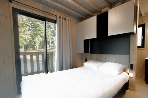 Modern ecolodge bedroom with large bed, overhead cabinets, and forest view through sliding glass door in Belgium.