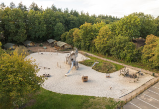 Parque infantil con toboganes y tiendas safari al fondo, rodeado de árboles y naturaleza, vista aérea.