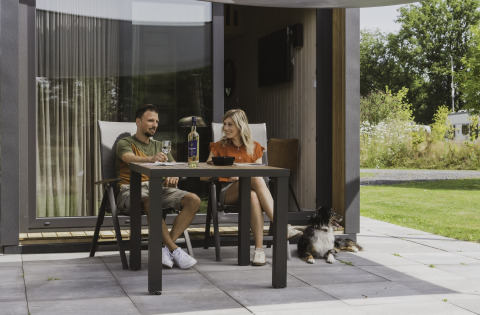 Couple assis devant une tiny house, partageant du vin à une table avec leur chien allongé sur la terrasse.