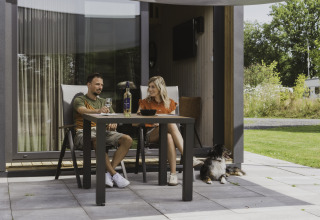 Pareja sentada fuera de una tiny house, disfruta de vino en la mesa y su perro descansa en la terraza.