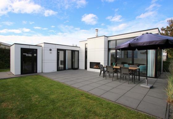 Lodge moderne L-Cube 6 avec grande terrasse, table extérieure, parasol et pelouse sous un ciel bleu.