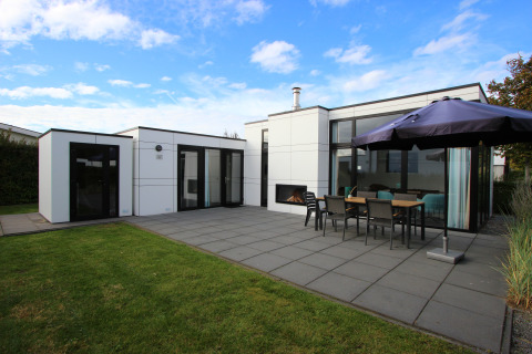Lodge moderne L-Cube 6 avec grande terrasse, table extérieure, parasol et pelouse sous un ciel bleu.