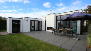 Lodge moderne L-Cube 6 avec grande terrasse, table extérieure, parasol et pelouse sous un ciel bleu.