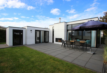 Moderne L-Cube 6 lodge met ruime patio, buiteneettafel, parasol en grasveld onder blauwe hemel.