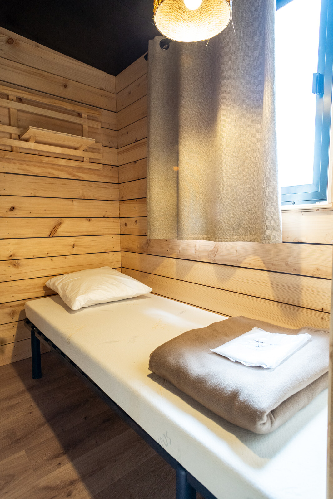 Eenvoudige houten cabine met eenpersoonsbed, kussen en raam in Evasion Chalet bij Camping d'Aix en Provence.