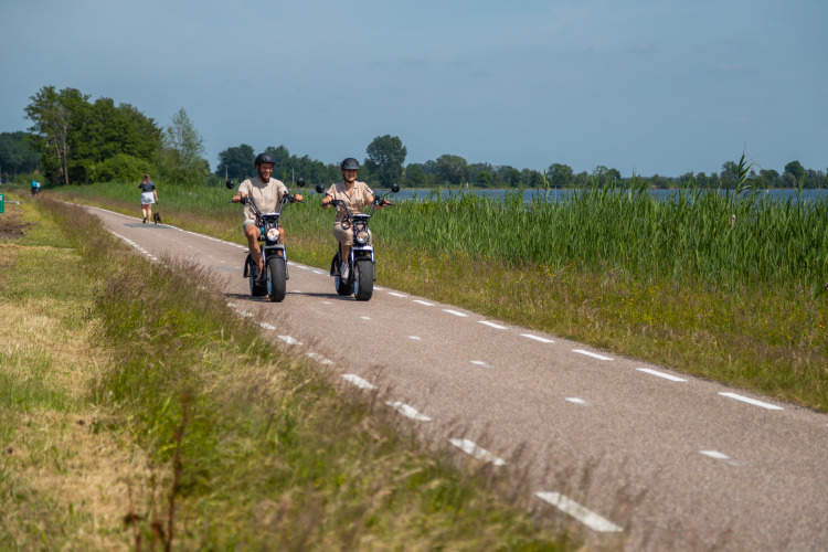 Zwei Personen fahren Elektroroller auf einem Radweg am See im Vakantiechalet comfort+ in den Niederlanden.
