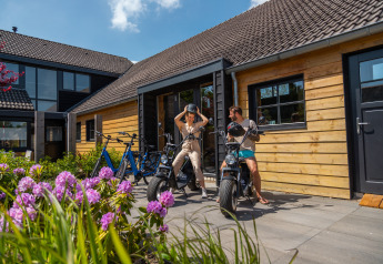 To personer gør sig klar til en cykeltur foran en træhytte med blomsterbed ved Vakantiepark De Wiedense Meren.