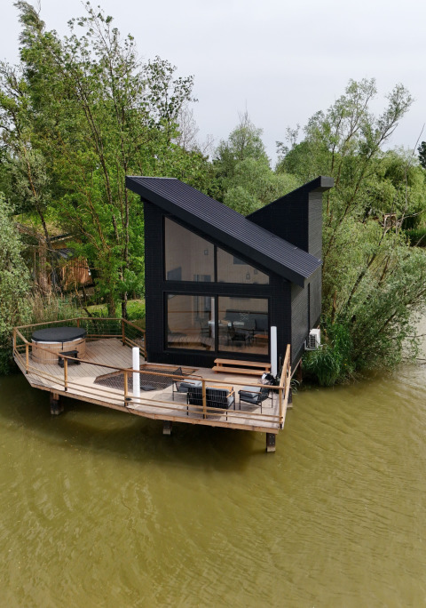Cabina moderna nera con tetto inclinato e terrazza sull’acqua al Domaine des Feuilles, Auvergne-Rhône-Alpes, Francia.