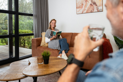 Kvinde læser en bog på sofaen i Vogellodge med en mand, der drikker vand, Holiday Resort 't Schuttenbelt.