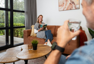 Frau liest ein Buch auf dem Sofa in der Vogellodge, während ein Mann ein Glas Wasser hält, Schuttenbelt.