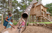 Family enjoying a playground with slide and wooden huts at Flower Camping Au Bois Des Biches, France.