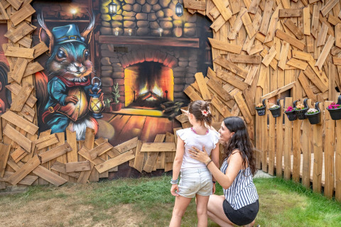 A woman and child admire a mural of a fairytale squirrel by a fireplace on a wooden wall at a holiday park.