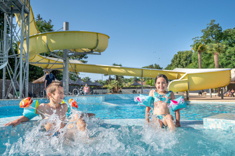 Twee kinderen met zwembandjes spelen in het zwembad bij een gele waterglijbaan in Flower Camping Au Bois Des Biches, Pays de la Loire.