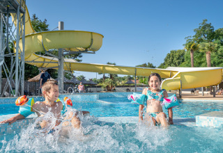 Zwei Kinder planschen mit Schwimmflügeln im Pool vor einer gelben Rutsche im Flower Camping Au Bois Des Biches, Pays de la Loire.