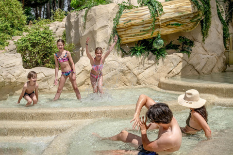 Børn og voksne nyder en tropisk swimmingpool med klipper og planter på Flower Camping Au Bois Des Biches.
