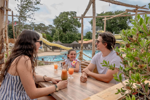 Une famille savoure des boissons à une table près de la piscine au Flower Camping Au Bois Des Biches, France.