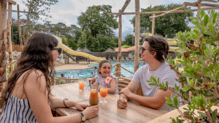 Eine Familie genießt Getränke an einem Tisch am Pool im Flower Camping Au Bois Des Biches, Frankreich.